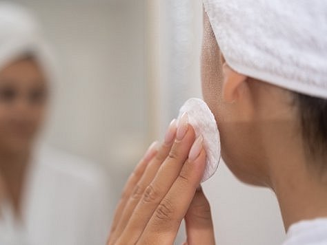 Persona frente al espejo usando un desmaquillante o agua micelar en un disco de algodón, limpiando el maquillaje de su cara. 